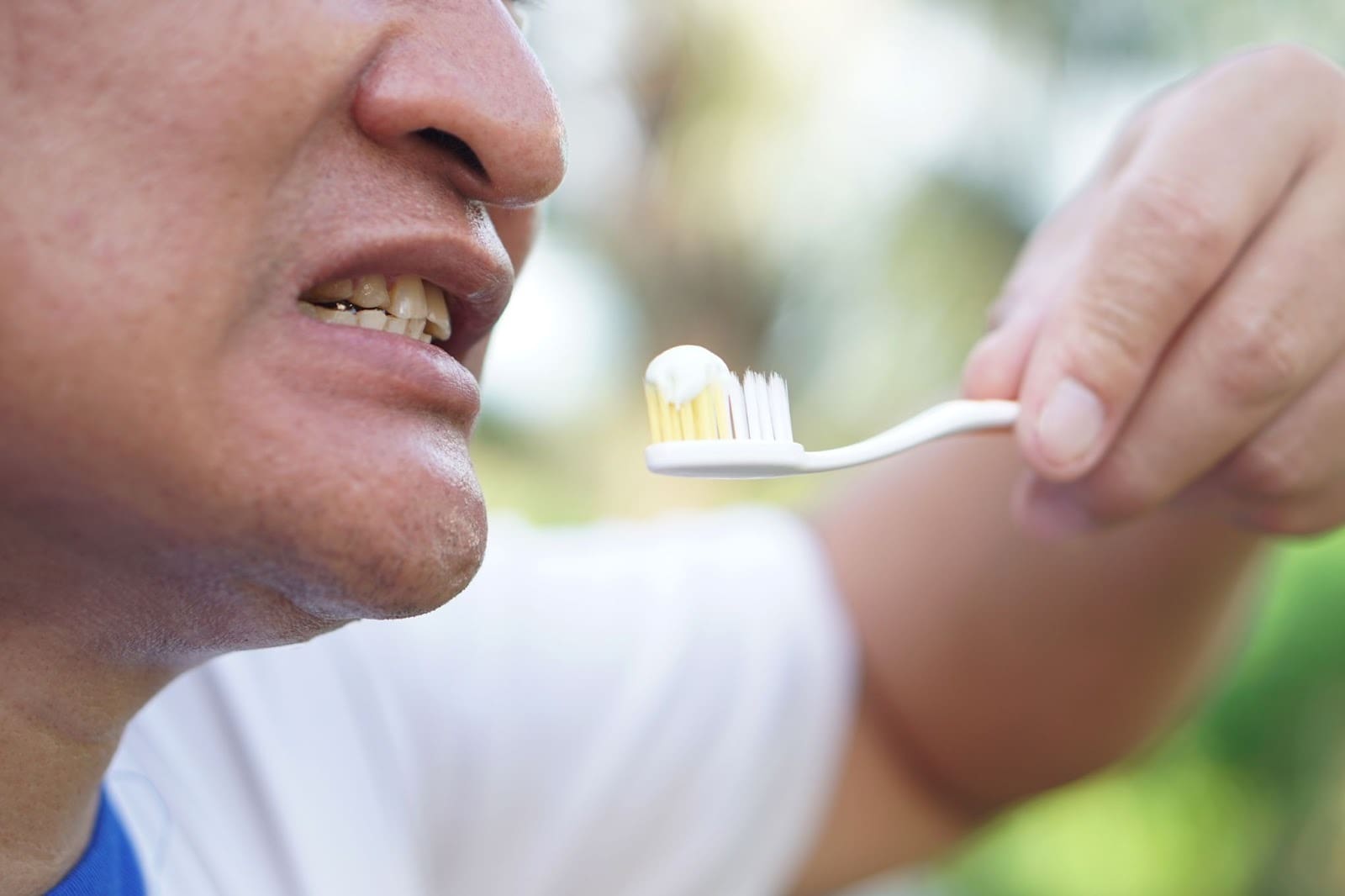 A close-up side profile of a man's mouth showing his teeth, while he holds a toothbrush with a small, pea-sized amount of toothpaste on the bristles.