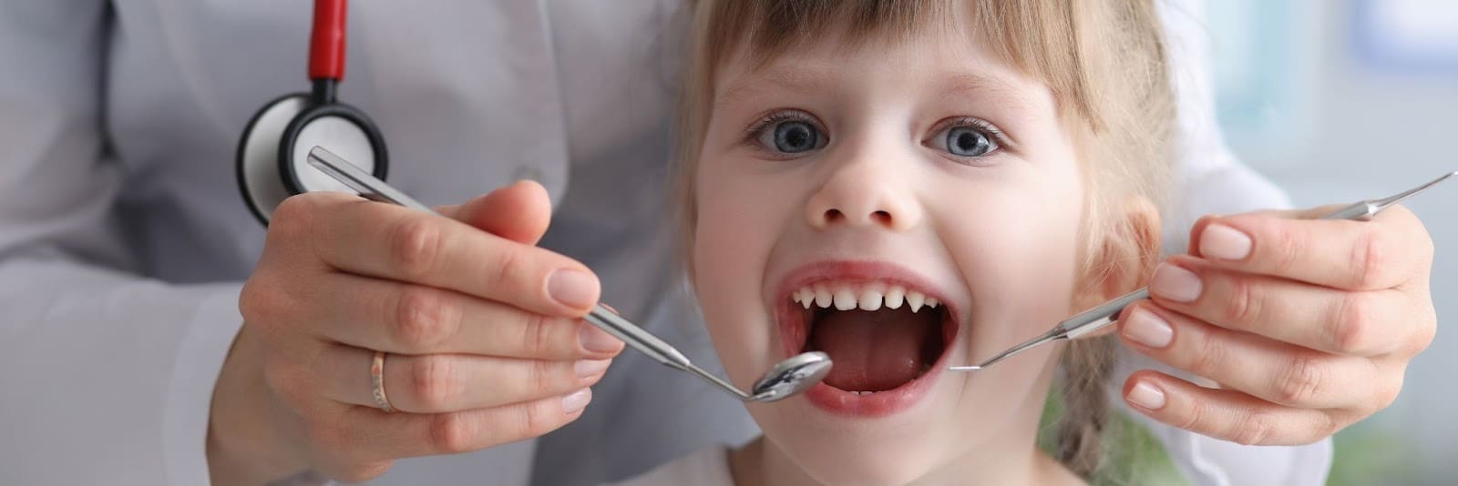 Close-up of a young child in a dental chair with their mouth open, while a pediatric dentist examines teeth with a mirror and explorer, emphasizing dentistry for kids.