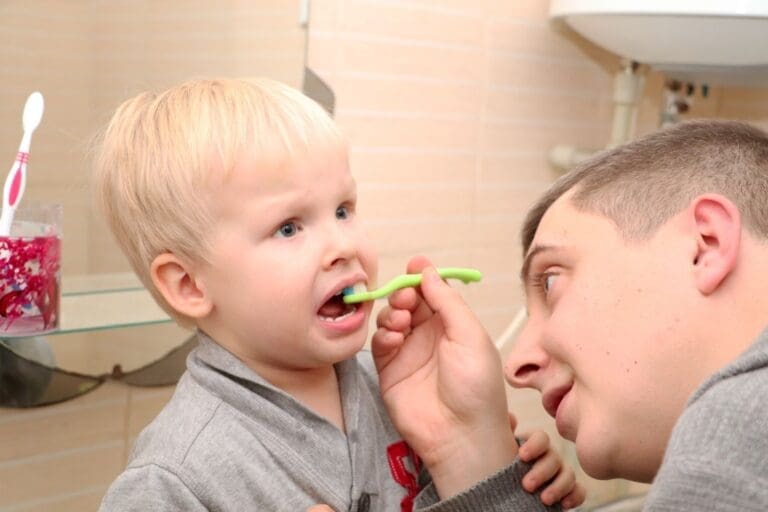 Close-up of a father in a gray top helping his young, blonde-haired son brush his teeth in a bathroom setting. The father gently holds the green toothbrush to the toddler's upper front teeth, while the boy looks up with a slightly resistant or concentrating expression. A cup with toothbrushes and red toothpaste residue is visible on a glass shelf in the background.