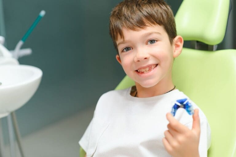 Close-up of a father helping his young, blonde-haired son brush his teeth in a bathroom. The father, seen from the side, holds a bright green toothbrush to the concentrating toddler's open mouth. The child wears a gray robe, and a mirror and toothbrushes in a cup are visible in the background.
