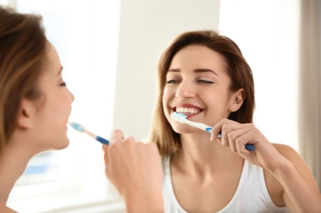 Happy woman smiling at her reflection while brushing her teeth, representing good dental hygiene services and a commitment to a clean, healthy smile.