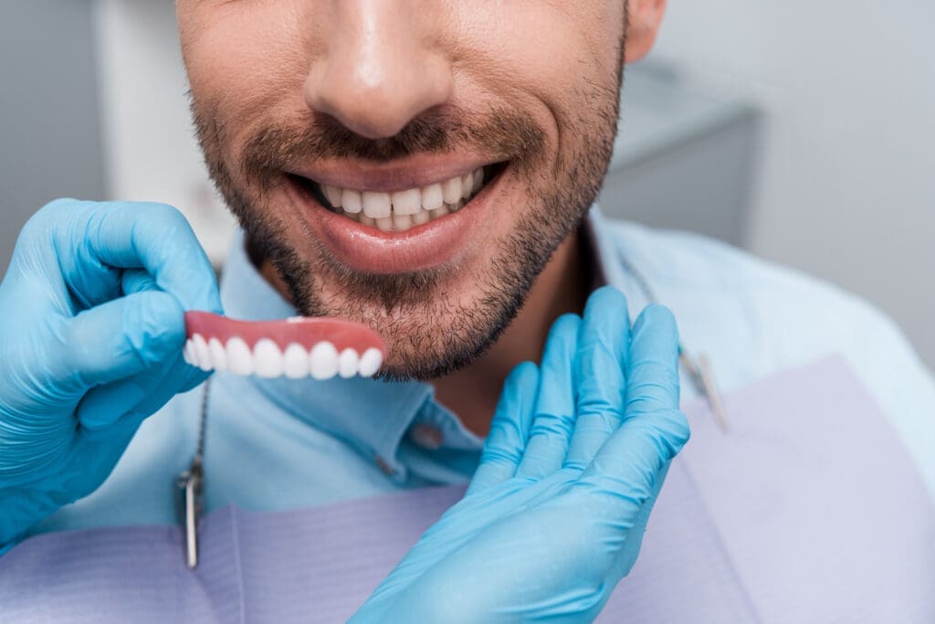 Close-up of a happy male patient smiling widely with his natural teeth visible, while a dental professional wearing blue protective gloves holds up a model of an upper dental prosthetic (denture or partial), illustrating a successful cosmetic or restorative fitting.