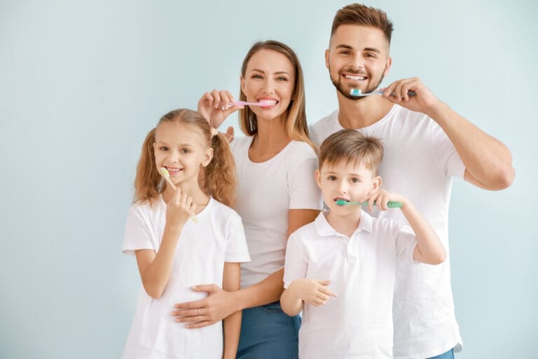 Happy Caucasian family—mother, father, daughter, and son—all wearing white shirts and jeans, standing together holding toothbrushes with a light blue background. The parents are smiling at the camera while brushing, representing family dental health and morning routine.