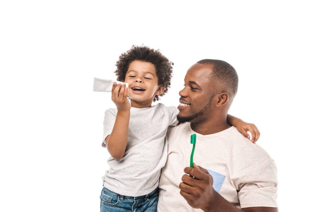 Smiling African American father and young son preparing to brush teeth, with the child holding toothpaste and the dad holding a green toothbrush, isolated on a white background. Represents family dental care, oral hygiene, and parenting.