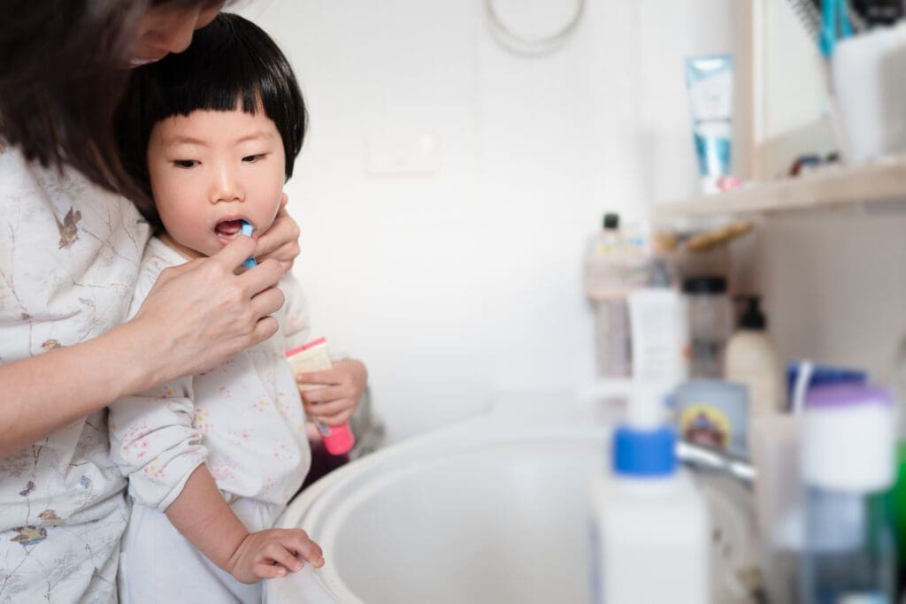 A mother in a bathroom is gently holding a toothbrush to her Asian toddler's mouth, demonstrating how to brush. The child is holding a pink tube of toothpaste while looking intently at the toothbrushing process over the sink. Focuses on early childhood dental care and oral hygiene routine.