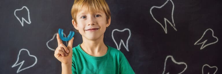 A smiling young boy in a green shirt holds up a bright blue athletic mouthguard in front of a dark chalkboard background decorated with chalk drawings of teeth, symbolizing child dental protection, sports safety, and preventative care.