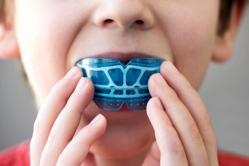 A cheerful young boy with a turquoise shirt smiles while holding a clear, customized dental appliance, likely a retainer or night guard, near his mouth against a light gray background, illustrating orthodontic treatment compliance or teeth grinding prevention.