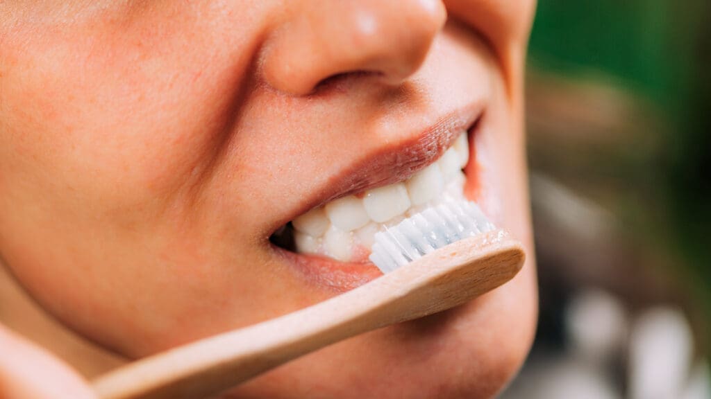 Close-up photo of a woman's mouth and white teeth as she brushes them with a sustainable, wooden bamboo toothbrush. Focuses on proper dental hygiene, natural oral care, and clean teeth.