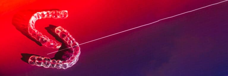 Clear, invisible dental aligners (braces/trays) and a single strand of white dental floss resting on a dramatic, gradient surface of red and dark blue/purple light. Used for invisible orthodontics and oral hygiene concepts.