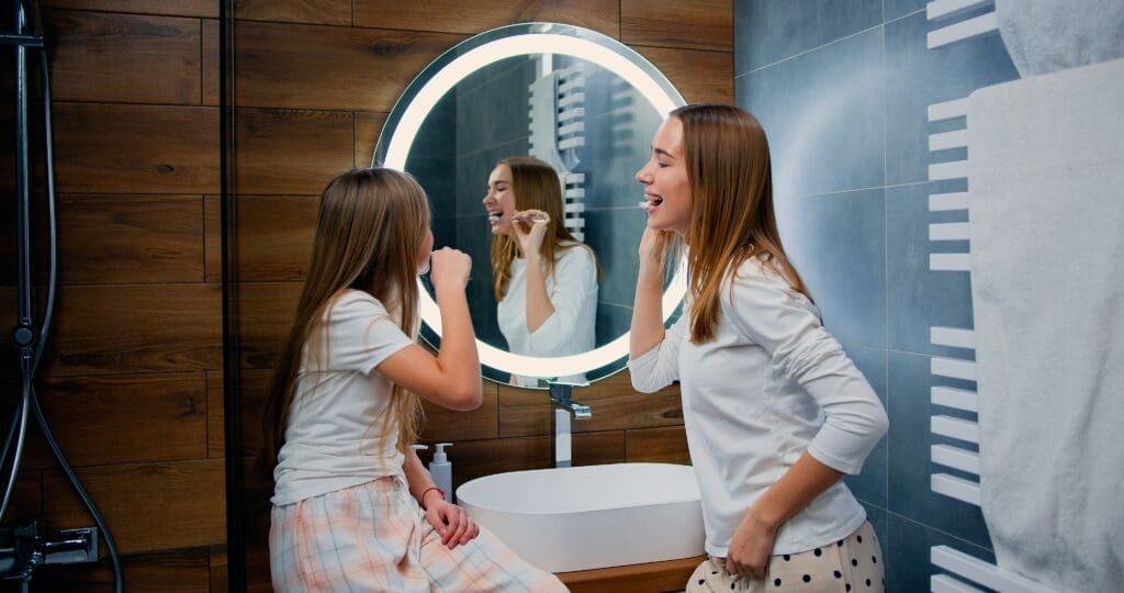 Two smiling girls, a mother and her young daughter, wearing pajamas and happily brushing their teeth together in a modern bathroom with a circular backlit mirror. Image represents family dental routines, teaching kids oral hygiene, and healthy habits.