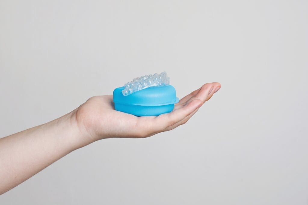 Close-up of a person's hand holding a clear plastic invisible aligner (orthodontic retainer) resting on a bright blue storage case, set against a light grey background. Image represents modern teeth straightening and dental health.