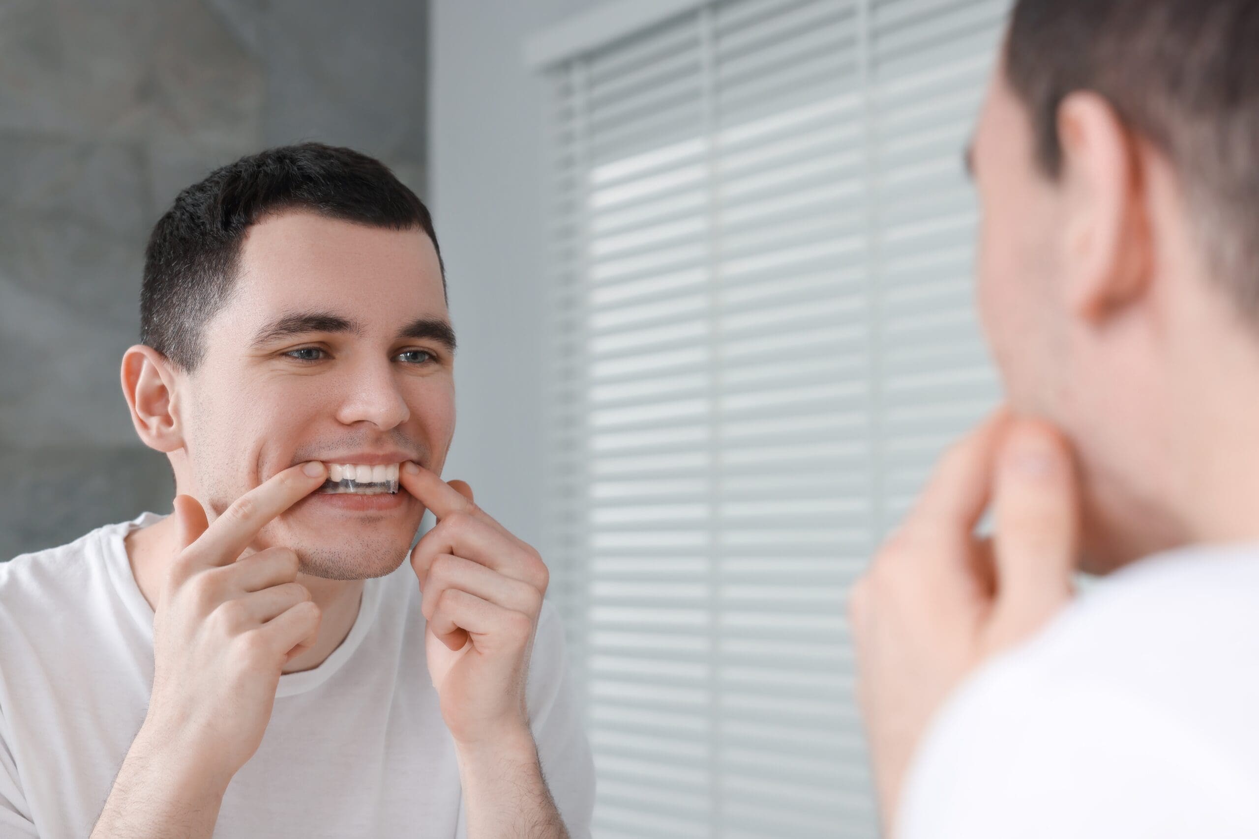 Young man in a white t-shirt checking his teeth in the bathroom mirror while wearing clear dental aligners, invisible braces, or a teeth whitening tray. Focus on cosmetic dentistry, adult orthodontics, and smile correction.