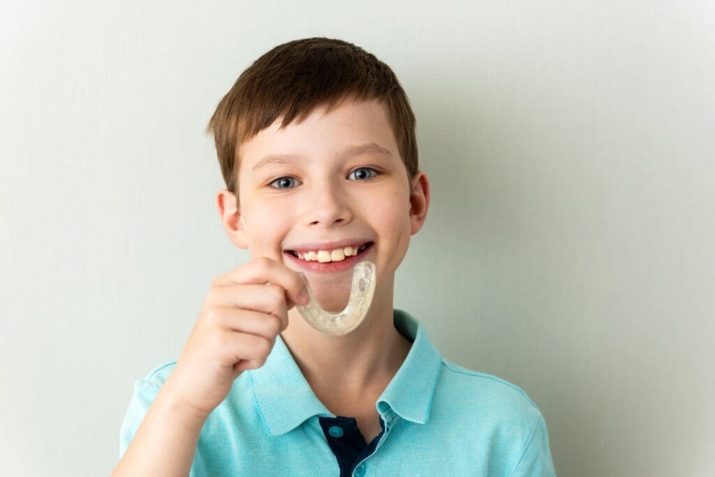 A smiling young boy in a green shirt holds up a bright blue athletic mouthguard in front of a dark chalkboard background decorated with chalk drawings of teeth, symbolizing child dental protection, sports safety, and preventative care.
