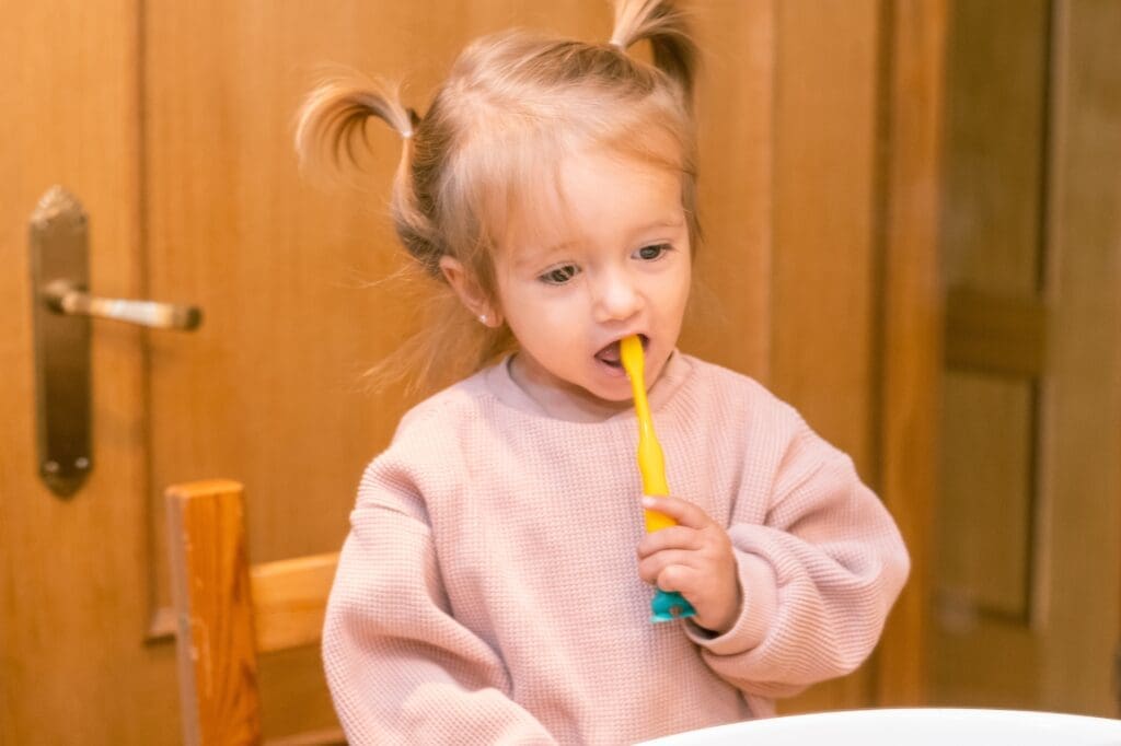 High-angle shot of a smiling baby lying on a couch while an adult gently lifts the upper lip to examine erupting front teeth. This image emphasizes the importance of a baby's first dental checkup and infant dental care.