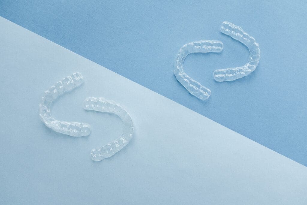 Two pairs of clear plastic orthodontic aligners resting on a geometric background split between light blue and white, representing discreet teeth straightening.